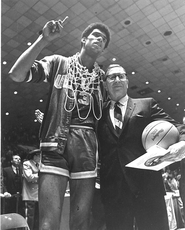 Lew Alcindor wears a UCLA number 33 jersey and a basketball net beside coach John Wooden in an archival arena photograph
