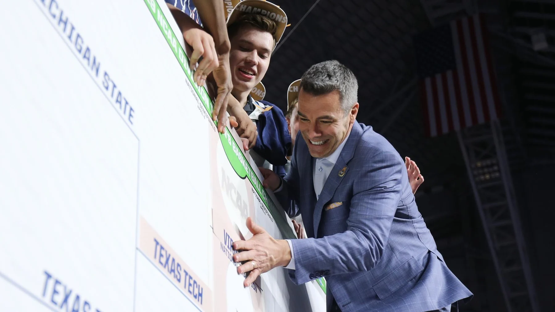 A coach in a blue suit and players gather around a large championship bracket board