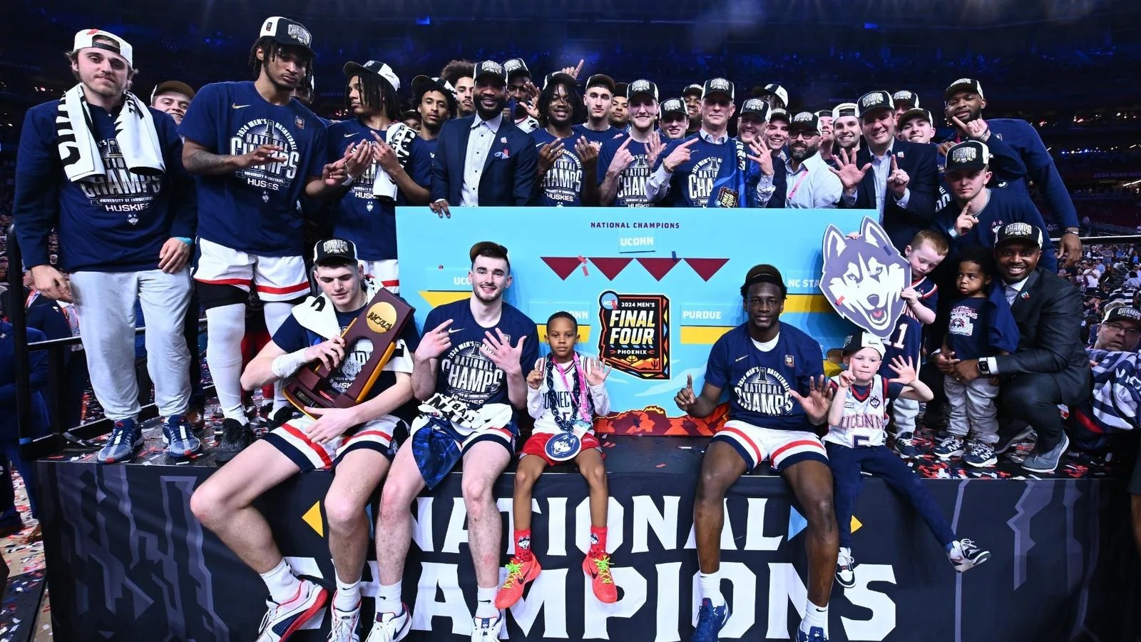 UConn players and staff posing with a national champions board and trophy on the Final Four stage