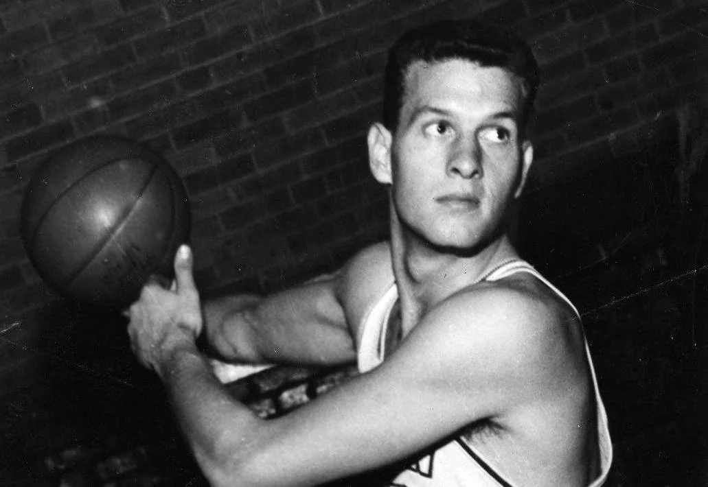 Frank Selvy in an archival basketball uniform holds a ball with both hands against a brick-wall background